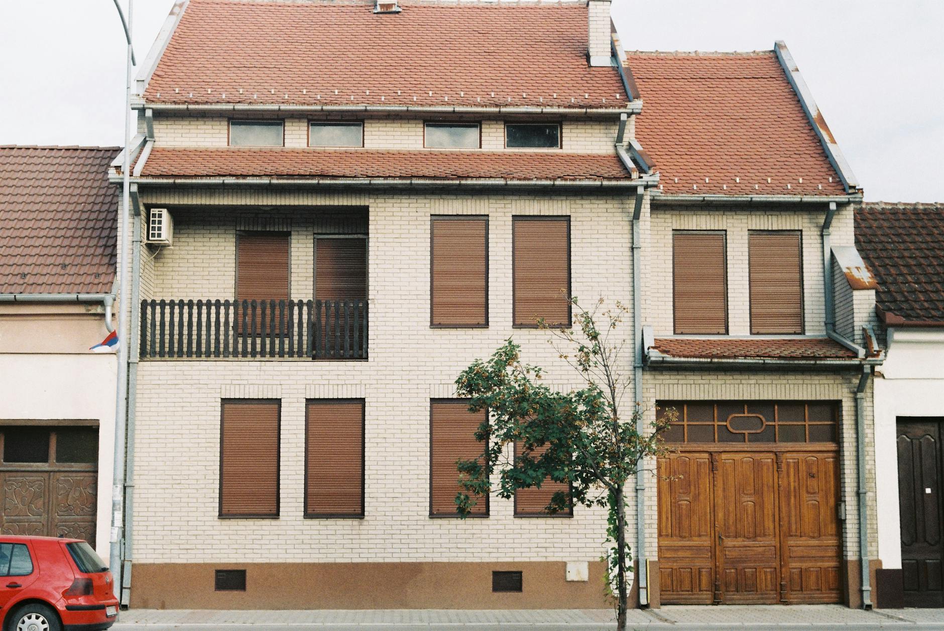 Brick house with brown shutters and garage, featuring a tree in front on a quiet street.